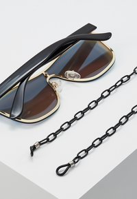 Saulesbrilles ar melniem rāmjiem, zelta akcentiem un brūnām gradienta lēcām, kopā ar melnu ķēdi ar saistītām sekcijām un metāla sprādzēm.