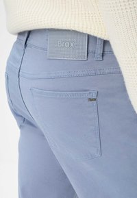 Pantaloni di cotone azzurri chiari con etichetta grigia texturizzata, cuciture decorative e una tasca posteriore con un piccolo marchio.