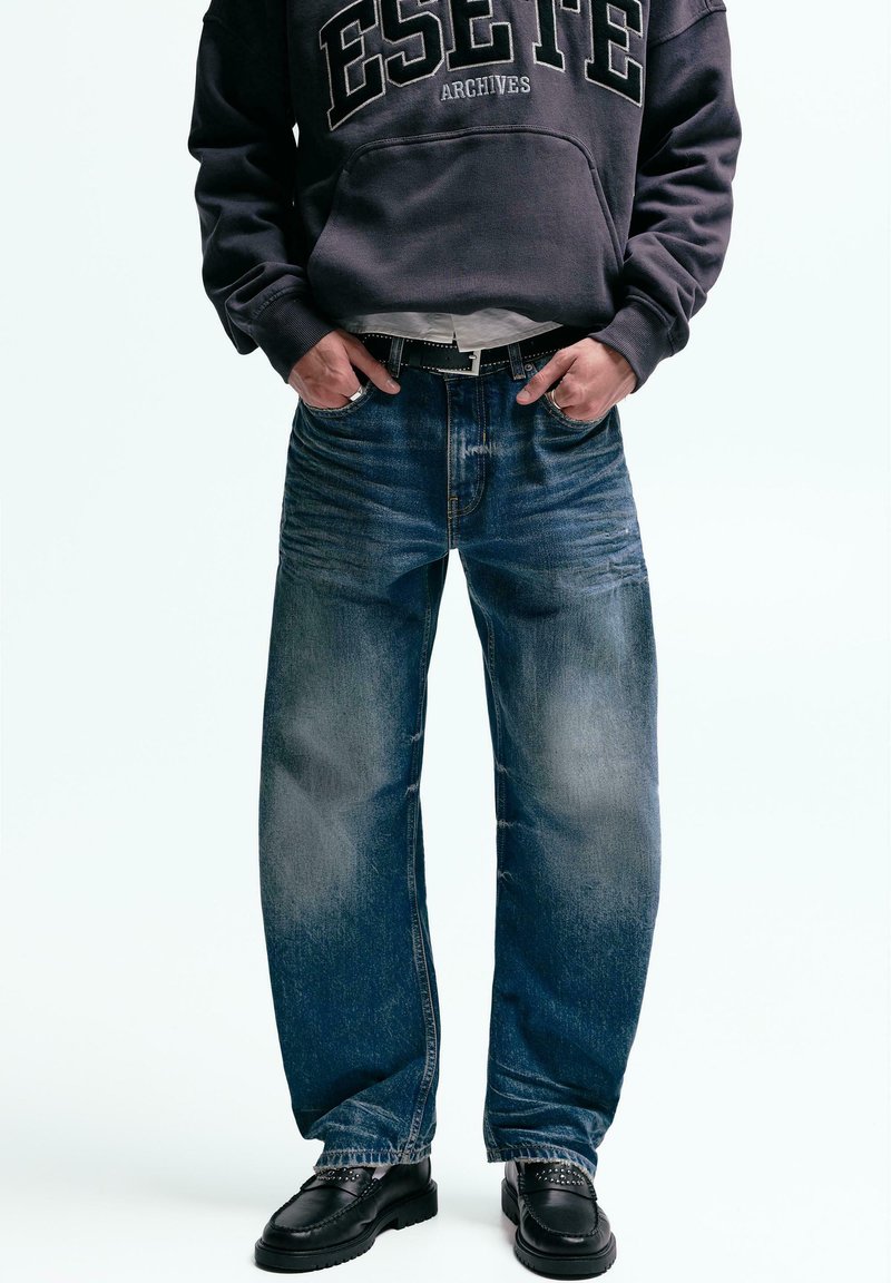 Jean en denim à coupe décontractée, avec un léger délavage et un design classique à cinq poches, assorti à un sweat-shirt gris et des chaussures noires.