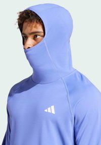 Junger Mann trägt ein helllila Balaclava-ähnliches Kapuzenshirt mit einem weißen dreieckigen Logo auf der Brust und blickt zur Seite.