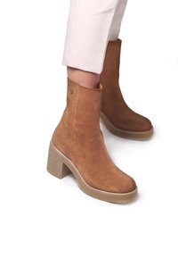 Brązowe zamszowe botki za kostkę z grubym obcasem, okrągłym noskiem i detalem w postaci guzika. Cholewka ma gładką teksturę i prosty design.