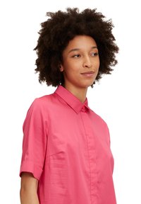 Kurzarm, rosa Hemd mit Knöpfen und Kragen sowie hochgekrempelten Ärmeln. Aus glattem Stoff mit glänzender Optik gefertigt.