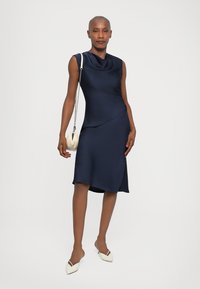 Rochie albastru marin cu decolteu tip cowl, design fără mâneci, tiv asimetric, textura netedă. Modelul ține o geantă mică de culoare crem.