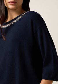Pull en maille bleu marine avec des manches larges et un décolleté arrondi. Accentué de strass transparents de différentes tailles le long du col.