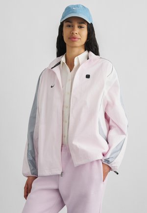 Vrouw draagt een lichtroze en grijze jas over een wit shirt, roze broek en een lichtblauwe Nike pet, staand met één hand in haar zak.
