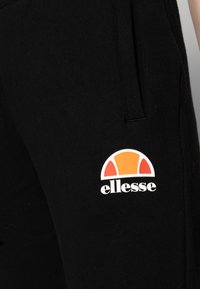 Svarta sweatpants med en vit och orange halvcirkel-logotyp med texten "ellesse". Tillverkade av mjukt material med sidofickor.