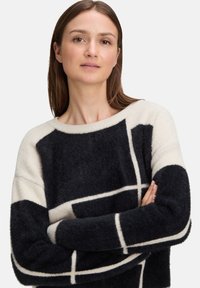 Zwart en crèmekleurig oversized trui met een fuzzy textuur, wijde halslijn en geometrisch patroon met gedurfde vierkanten en strepen op de mouwen.