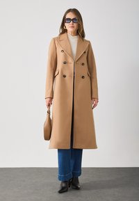 Lang kamelkleurige trenchcoat met een dubbelgebreid ontwerp, zwarte knopen, klepzakken, gecombineerd met blauwe flared jeans en zwarte enkellaarzen.