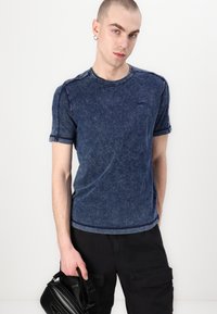 Tricou bleumarin, cu mânecă scurtă, cu o textură de denim decolorat, guler rotund și detalii de cusătură contrastante. Purtați-l cu pantaloni cargo negri.