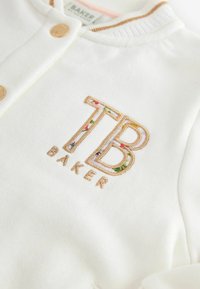 Weißer Pullover mit goldenen Druckknöpfen, geripptem Kragen und gesticktem "TB BAKER"-Logo mit floralen Details in Pink, Grün und Blau.