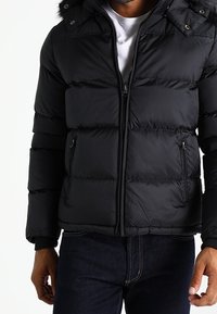 Black puffer jacket with a zip front, two side pockets, and a detachable hood. Features horizontal quilted design and snap buttons.