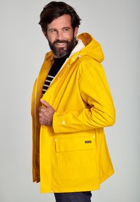 Gul vandtæt regnjakke med hætte, front snap-lukninger, en stor ydre lomme og hvidt for. Designet med en afslappet pasform.
