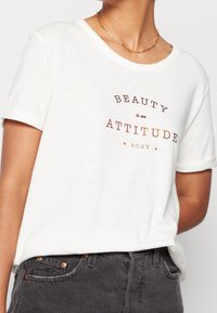 Weißes T-Shirt mit kurzen Ärmeln, das braunen Text mit der Aufschrift "SCHÖNHEIT ist eine EINSTELLUNG" zeigt, darunter "ROXY". Aus weichem, leichtem Stoff mit lockerer Passform.