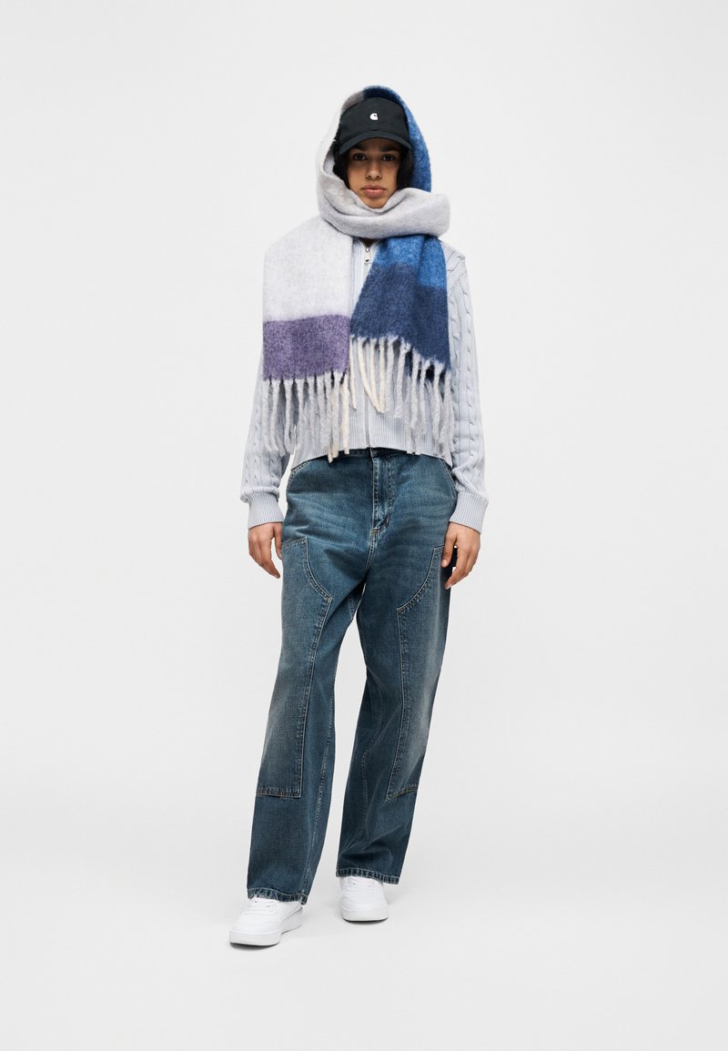 Personne portant une casquette noire, une grande écharpe bleue et violette, un pull en laine bleu clair, un jean large en denim et des baskets blanches, debout sur un fond gris.