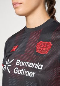 Čierny futbalový dres s červenými akcentmi, okrúhlym výstrihom a štruktúrovaným vzorom. Zobrazuje logá Bayeru Leverkusen a Barmenia Gothaer.