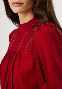 Blusa rossa con collo alto, tessuto testurizzato, design plissettato e cuciture decorative a catena lungo il busto.
