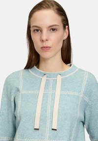 Himmelblauer Strickpullover mit strukturiertem Muster, runder Ausschnitt und beigen Kordeln mit metallischen Akzenten.