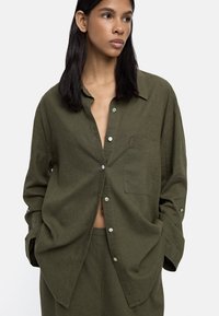 Kobieta z długimi ciemnymi włosami, ubrana w oliwkową zieloną koszulę na guziki i dopasowane spodnie, z rękami w kieszeniach, patrząca w bok na tle białego tła.