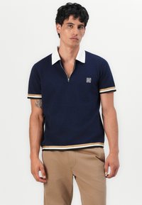 Muška tamnoplava polo majica na zip s bijelim ovratnikom, zlatnim i crnim prugama, teksturiranom tkaninom i vezom logotipa na prsima.