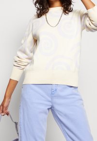 Cremfarbener Pullover mit helllila Spiralmustern, gerippten Bündchen und Saum. Kombiniert mit hellblauen Hosen, die einen lockeren Sitz und einen lässigen Look zeigen.