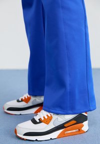 Nike Air Max tenisice u bijeloj, sivoj i crnoj boji s narančastim naglascima. Plave hlače imaju glatku teksturu i ravni kroj, dosežući sredinu tele.