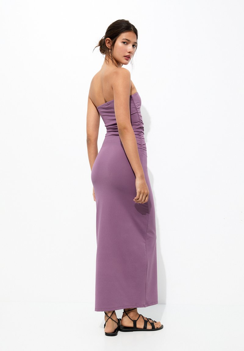 Robe maxi sans bretelles violet avec des détails froncés le long du buste, texture lisse et silhouette ajustée, associée à des sandales noires.