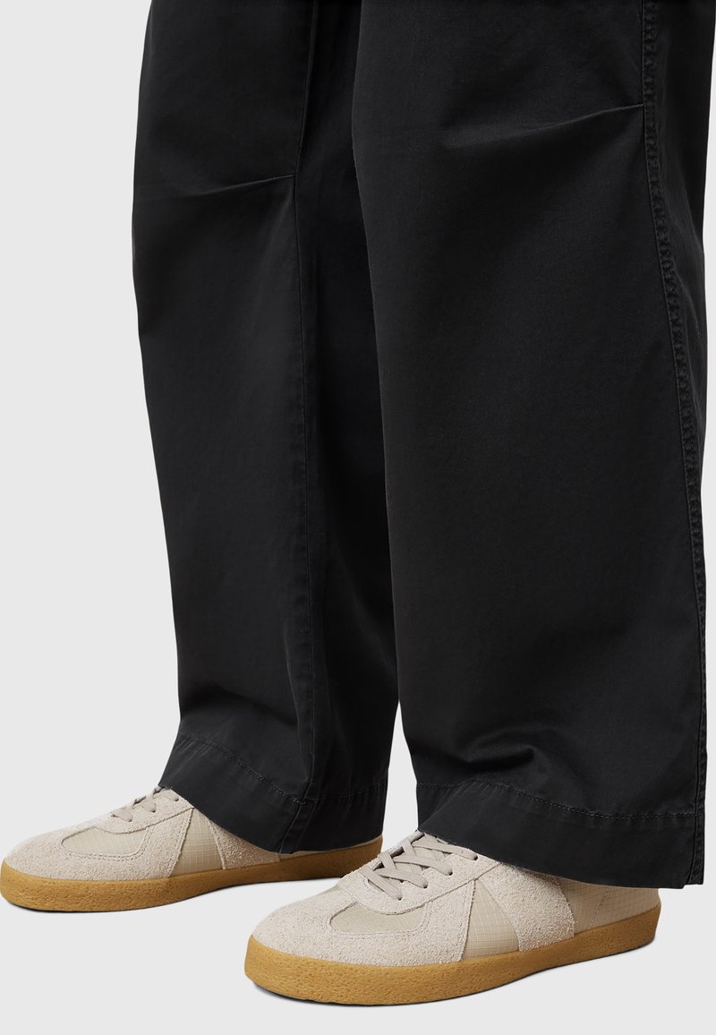 Beige Textursneaker mit einer Gummisohle, ausgestattet mit leichtem Wildleder, Mesh-Akzenten und einem gepolsterten Kragen, kombiniert mit lockeren schwarzen Hosen.