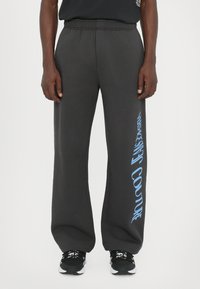 Black sweatpants with an elastic waistband, featuring a blue graphic print of "Versace Jeans Couture" on the left leg. Material is soft and textured.