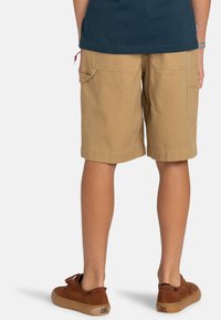 Shorts cargo beiges avec poches latérales, coupe droite, ourlets retroussés, fabriqués en tissu en coton durable. Portés avec des chaussures marron.