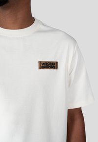 Biała bawełniana koszulka z okrągłym dekoltem, z brązową metką z tkaniny, na której czarną nicią wyszyto "WRONG FRIENDS". Prosty, schludny design.