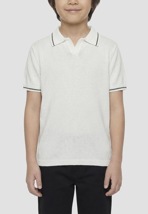 Kind draagt een wit poloshirt met korte mouwen met donkere rand op de kraag en mouwen, gecombineerd met zwarte broek, staand tegen een effen achtergrond.
