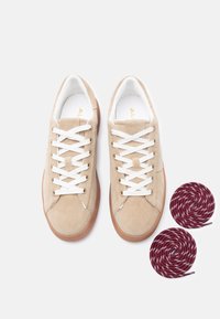 Baskets en daim beige avec des lacets blancs, des semelles en gomme, et deux lacets de rechange enroulés, bordeaux et blancs, à côté, sur un fond blanc.