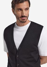 Schwarze Strickjacke mit V-Ausschnitt und fünf Knöpfen, getragen über einem weißen T-Shirt. Hat eine weiche Textur, eine schlanke Passform und ein minimalistisches Design.