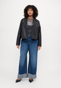 Veste de moto en cuir noir sur un haut gris à imprimé léopard, associée à un jean en denim bleu à jambes larges avec un ourlet effiloché. Des talons noirs complètent le look.
