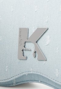 Svetlomodrá denimová čiapka s strieborným kovovým logom K, s viditeľnou textúrou a detailmi šitia, zdôrazňujúca voľný a neformálny dizajn.