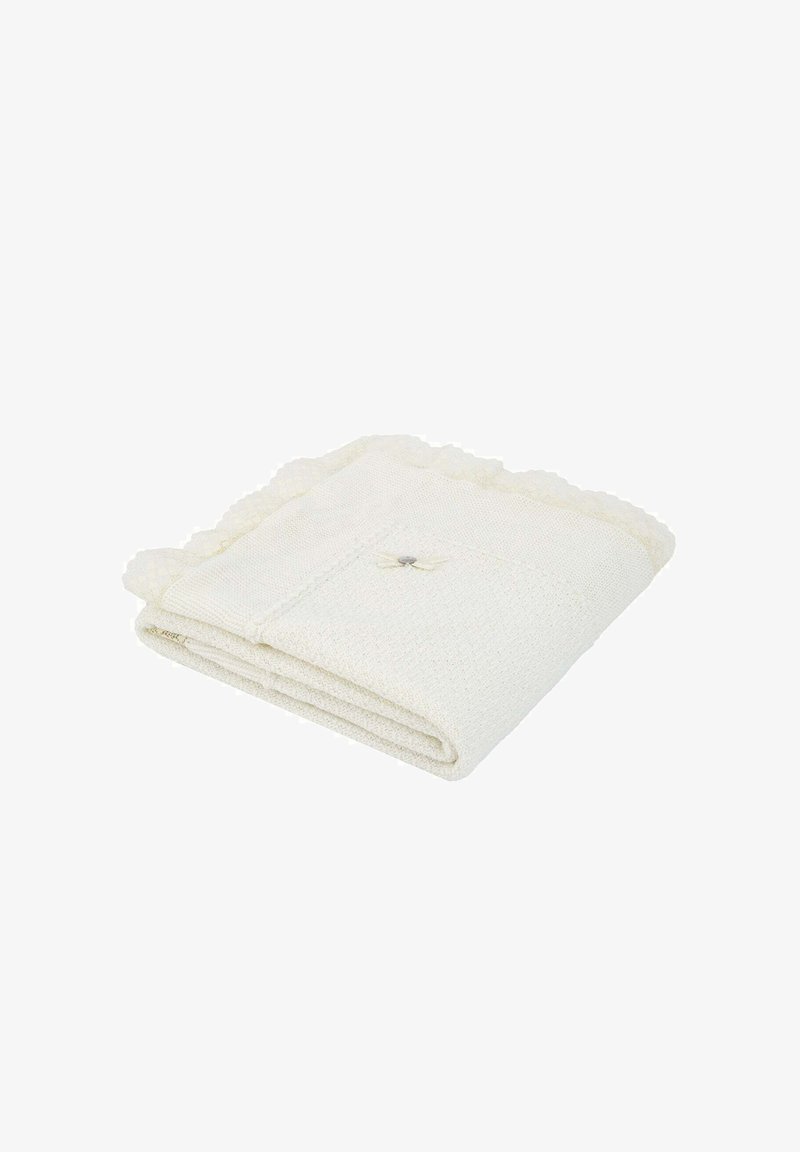 Couverture en tricot couleur crème, pliée, avec des bords en scallop et un petit nœud décoratif dans un coin.
