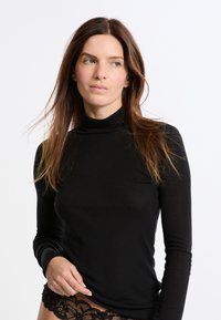 Donna con lunghi capelli castani che indossa un maglione nero a collo alto e biancheria intima in pizzo nera, che guarda di lato contro uno sfondo bianco.