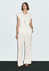 Haut blanc avec un design drapé et des manches courtes, associé à un pantalon ample de couleur claire. Les accessoires comprennent un long collier avec un pendentif en pierre.