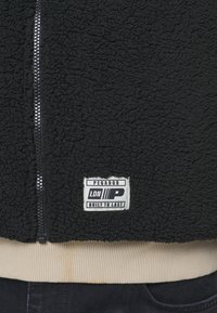 Czarna kurtka z polaru o fakturowanej powierzchni, z srebrnym zamkiem błyskawicznym i beżowym prążkowanym dołem. Posiada metkę z napisem "PEGADOR LDN BUILT TO LAST."