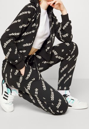 Osoba klęcząca, ubrana w czarny dres Adidas z białymi logo oraz białe buty sportowe Adidas z czarnymi paskami i turkusowymi akcentami.
