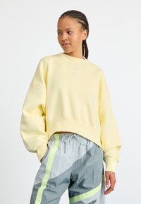 Giovane donna con capelli intrecciati, che indossa una felpa Nike gialla chiara e pantaloni da jogging grigi con strisce verde neon, che guarda di lato.