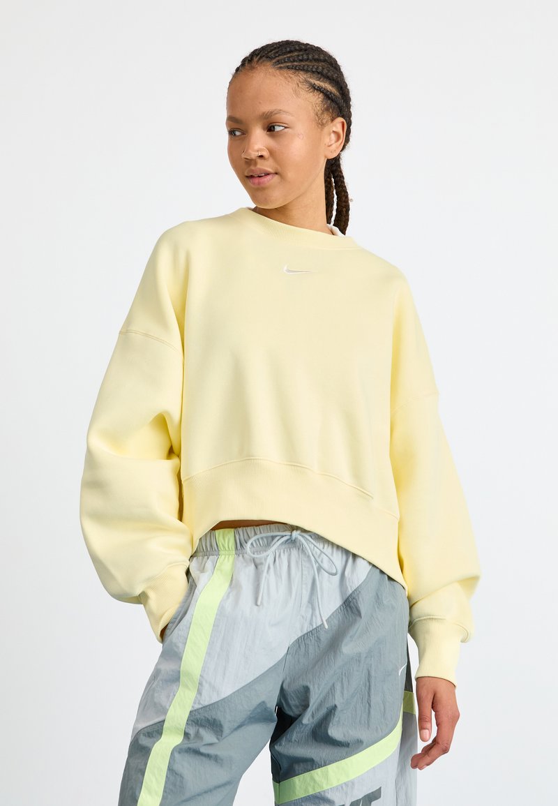 Giovane donna con capelli intrecciati, che indossa una felpa Nike gialla chiara e pantaloni da jogging grigi con strisce verde neon, che guarda di lato.