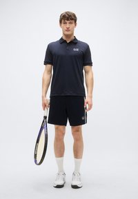 Tmavomodré polo tričko s krátkymi rukávmi, logo na hrudi, v kombinácii s čiernymi šortkami s logom, držiace tenisovú raketu, obuté v bielych ponožkách a topánkach.