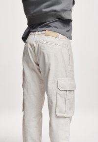 Beige corduroy cargo bukser med sidelommer og frontfolder, med et kvadratisk mærke på taljen mærket "2Y STUDIOS."