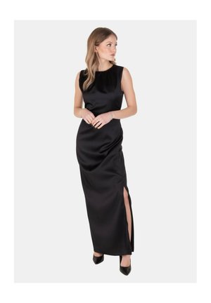 Donna in abito lungo nero in satin senza maniche con spacco laterale, indossa tacchi neri, guarda verso sinistra su sfondo bianco semplice.