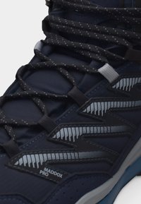 Lähikuva tummansinisen Maddox Pro -lenkkarin mustavalkokuvioisista nauhoista ja heijastavista sivuaksenteista.