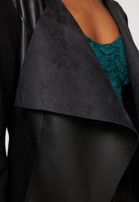 Schwarze Lederjacke mit einem weichen, dunkelgrauen Wildlederrevers und einem leuchtend türkisfarbenen Spitzentop darunter. Das Design hat eine taillierte Silhouette.