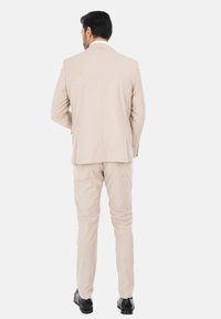 Abito beige realizzato in tessuto leggero, con giacca monopetto e pantaloni abbinati, completo di eleganti scarpe nere.