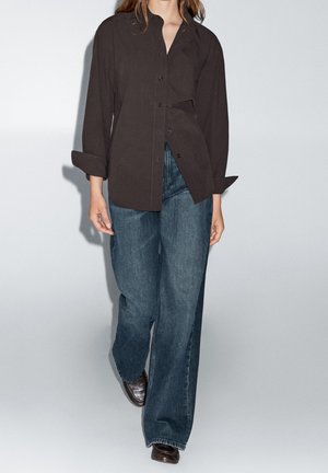 Woman wearing a loose dark brown button-up shirt, wide-leg blue jeans, and dark brown shoes, walking against a plain light background.