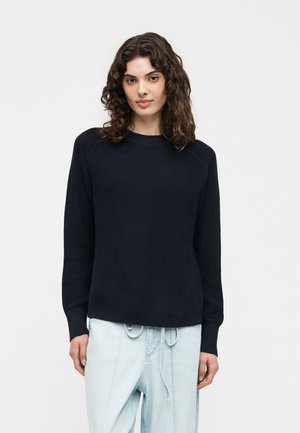 Femme aux cheveux bruns bouclés portant un pull en maille noir et un jean taille haute bleu clair, debout devant un fond blanc uni.